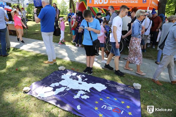 Na trawie leży baner z mapą Europy. Przed nim stoi chłopiec w niebieskiej koszulce i robi zdjęcie telefonem. Na drugim panie kilkanaście osób, stoi lub idzie oraz część pomarańczowego namiotu.