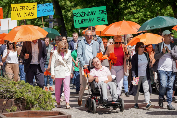 Grupa osób z niepełnosprawnościami razem z opiekunami idzie ulicą Kielc. Część osób trzyma pomarańczowe parasolki. Są też transparenty, jeden z napisem "Różnimy się, lubimy się".