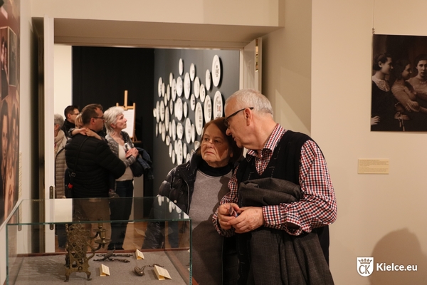 Kobieta i mężczyzna oglądają wystawę w Muzeum Historii Kielc. Na drugim planie ściana, wiszą na niej talerze. Przed ścianą stoi kilka osób.