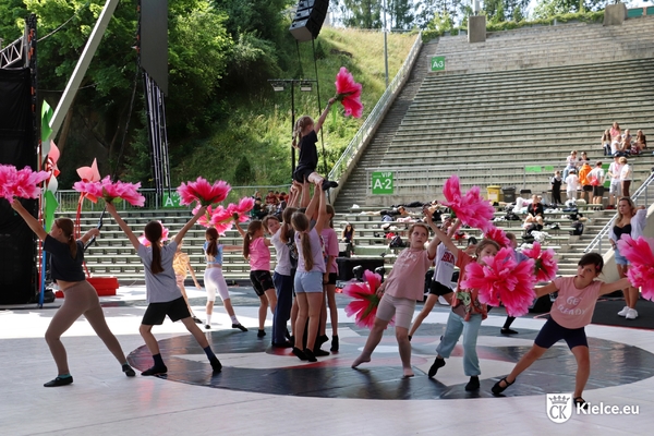 Grupa dzieci na scenie Amfiteatru Kadzielnia, tancerze trzymają różowe kwiaty z papieru, w tle widać miejsca dla publiczności