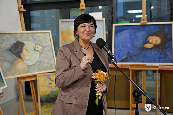 Autorka prac Agnieszka Łuczyńska stojąca przy mikrofonie, za nią ustawione są obrazy