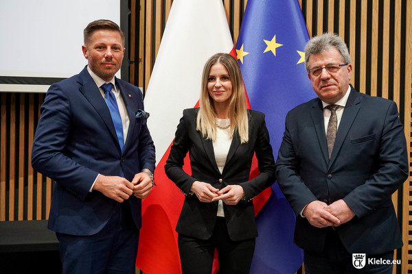 Kobieta i dwóch mężczyzn pozuje do zdjęcia. W tle flagi Polski i Unii Europejskiej.