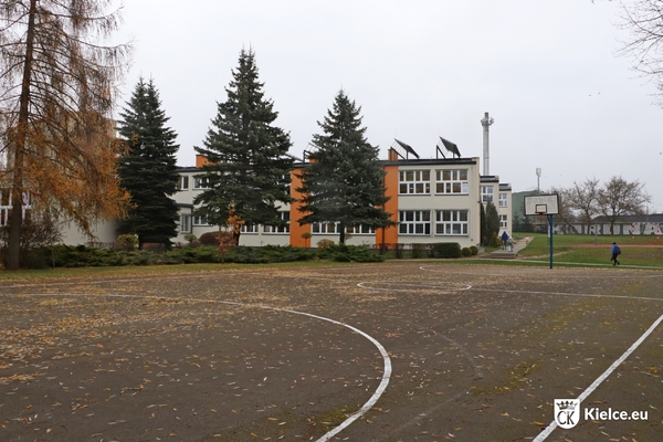 Na pierwszym planie boisko asfaltowe, na drugim planie drzewa i budynek szkoły oraz boisko trawiaste.