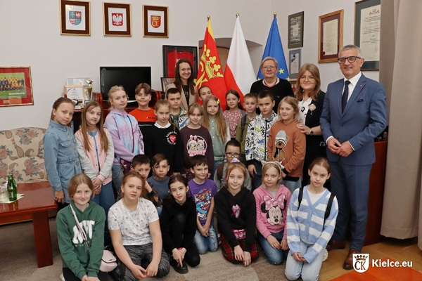 Kilkanaścioro dzieci w gabinecie prezydenta Kielc oraz cztery osoby dorosłe. W tle stoją flagi.