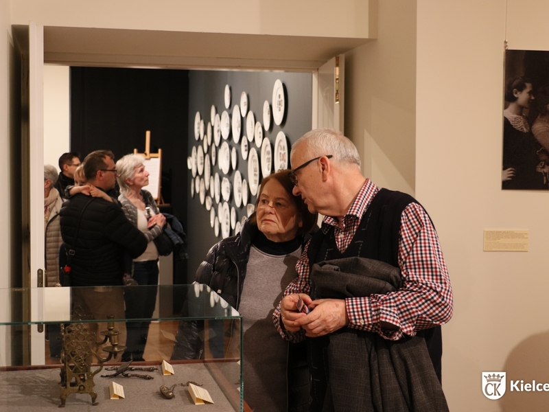 Kobieta i mężczyzna oglądają wystawę w Muzeum Historii Kielc. Na drugim planie ściana, wiszą na niej talerze. Przed ścianą stoi kilka osób.